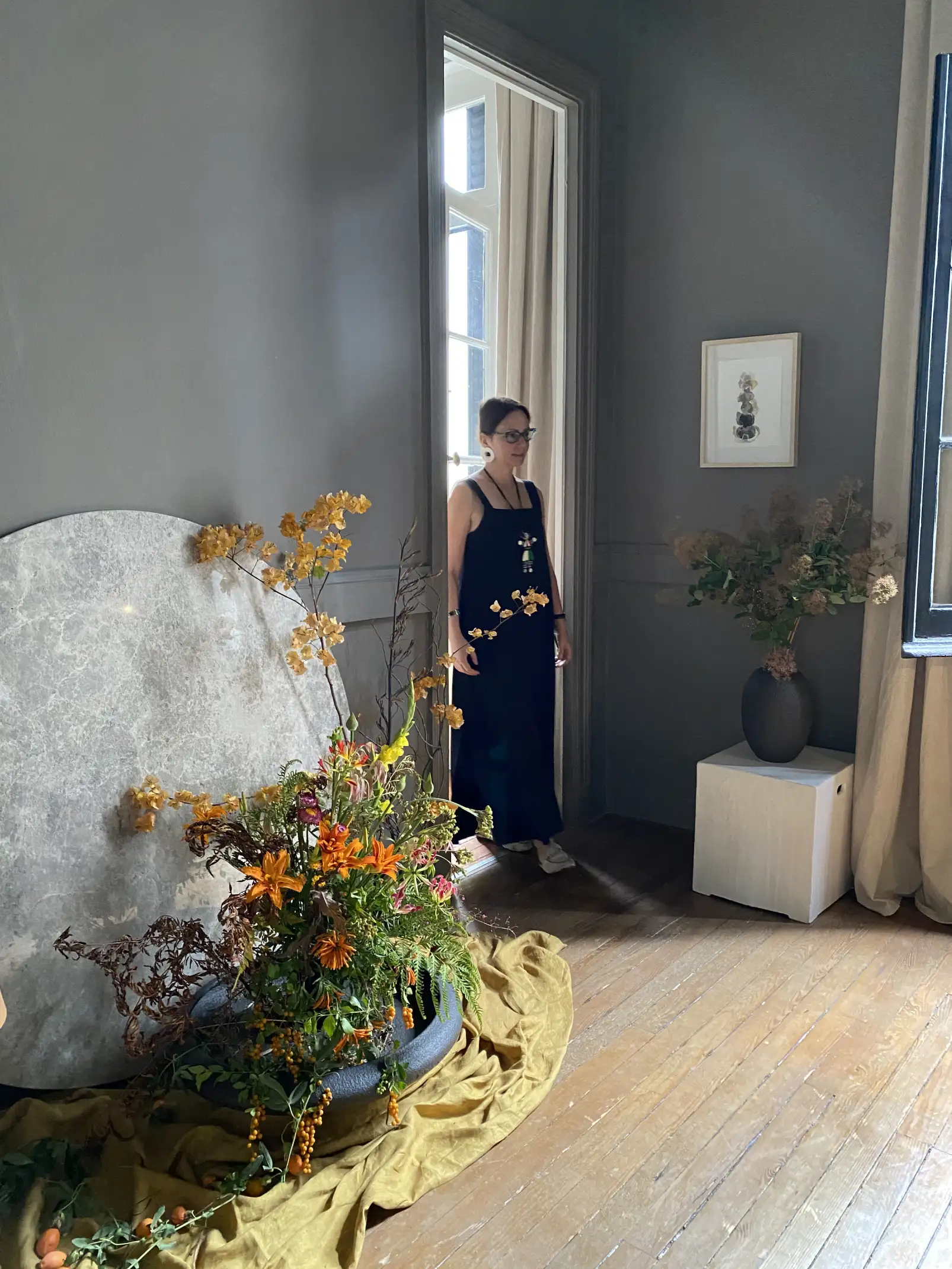 Irina Khatsernova en un interior luminoso junto a una instalación floral con lirios naranjas y follaje, en ambiente artístico y natural.