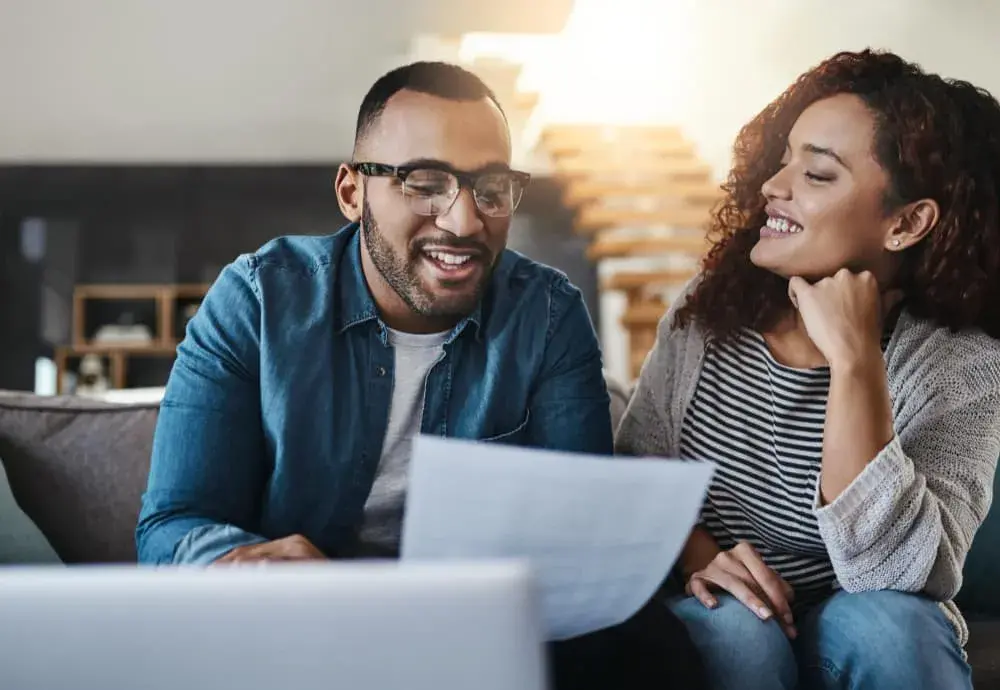 A smiling couple sits together reviewing financial documents on a couch with a laptop open in front of them. The image represents clarity and confidence in managing investment property financing through Debt Service Coverage Ratio (DSCR) loans from Chris Lewis Home Loans.