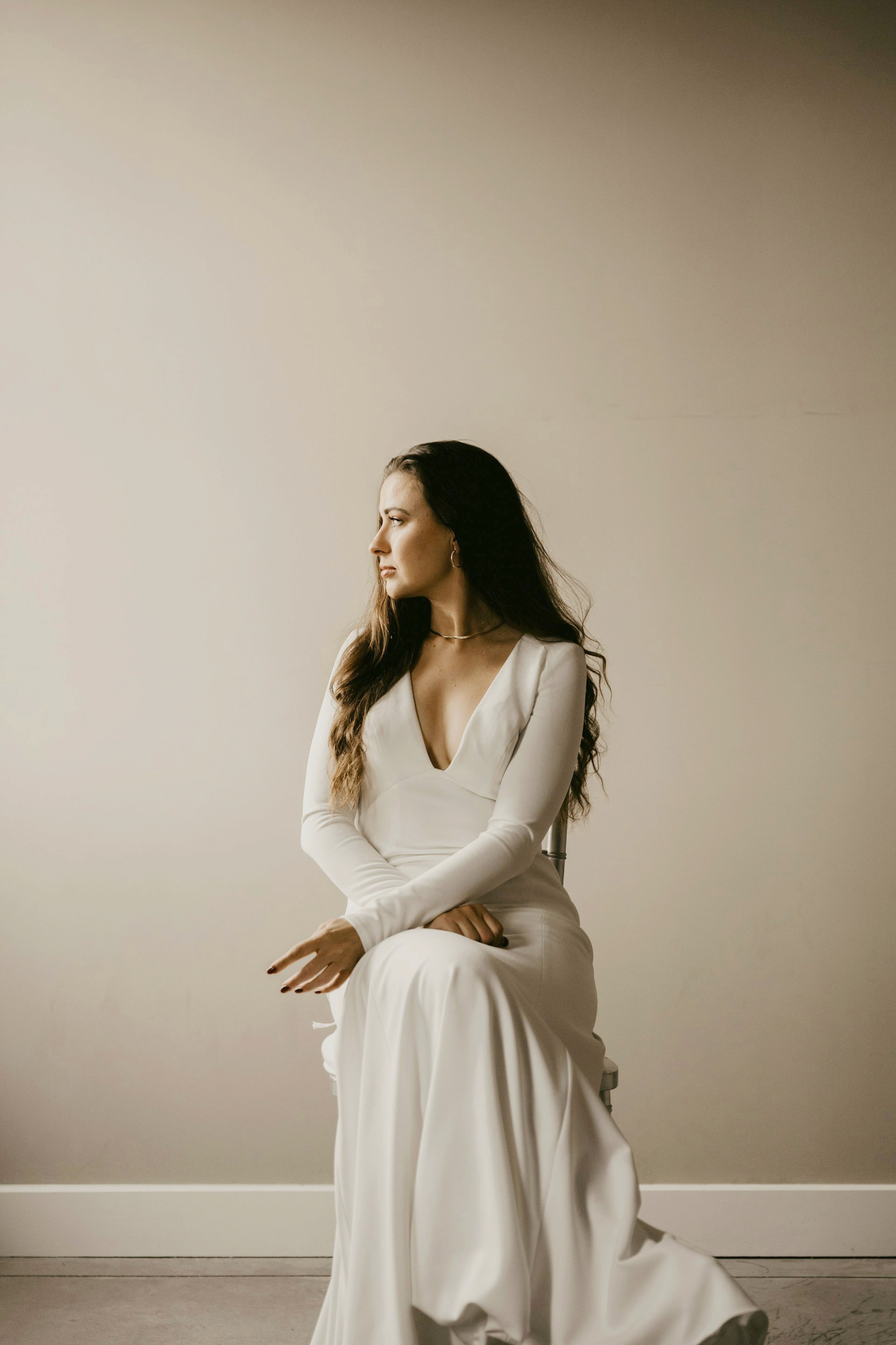 A woman in a long-sleeved, deep V-neck white gown sitting on a chair and looking away from the camera in a soft-lit room.