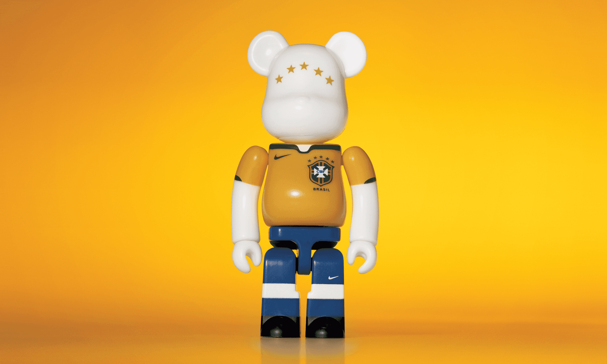 A single 3.5” tall Bre@rbrick/Nike toy created to commemorate the 2006 Brazilian World Cup football team. The toy is painted to appear in the Brazilian football kit and stands in the center of the frame in front of a bright yellow background.