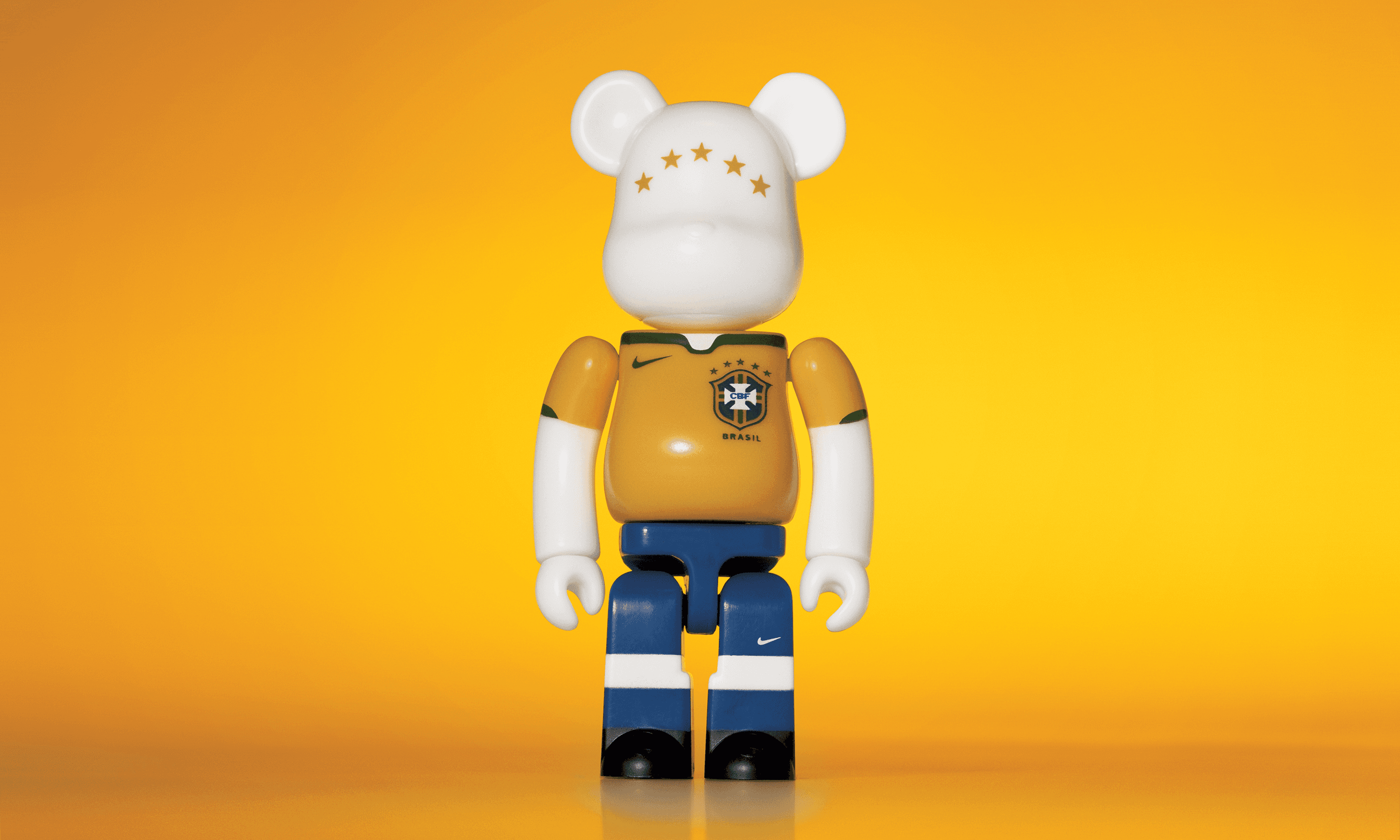 A single 3.5” tall Bre@rbrick/Nike toy created to commemorate the 2006 Brazilian World Cup football team. The toy is painted to appear in the Brazilian football kit and stands in the center of the frame in front of a bright yellow background. 