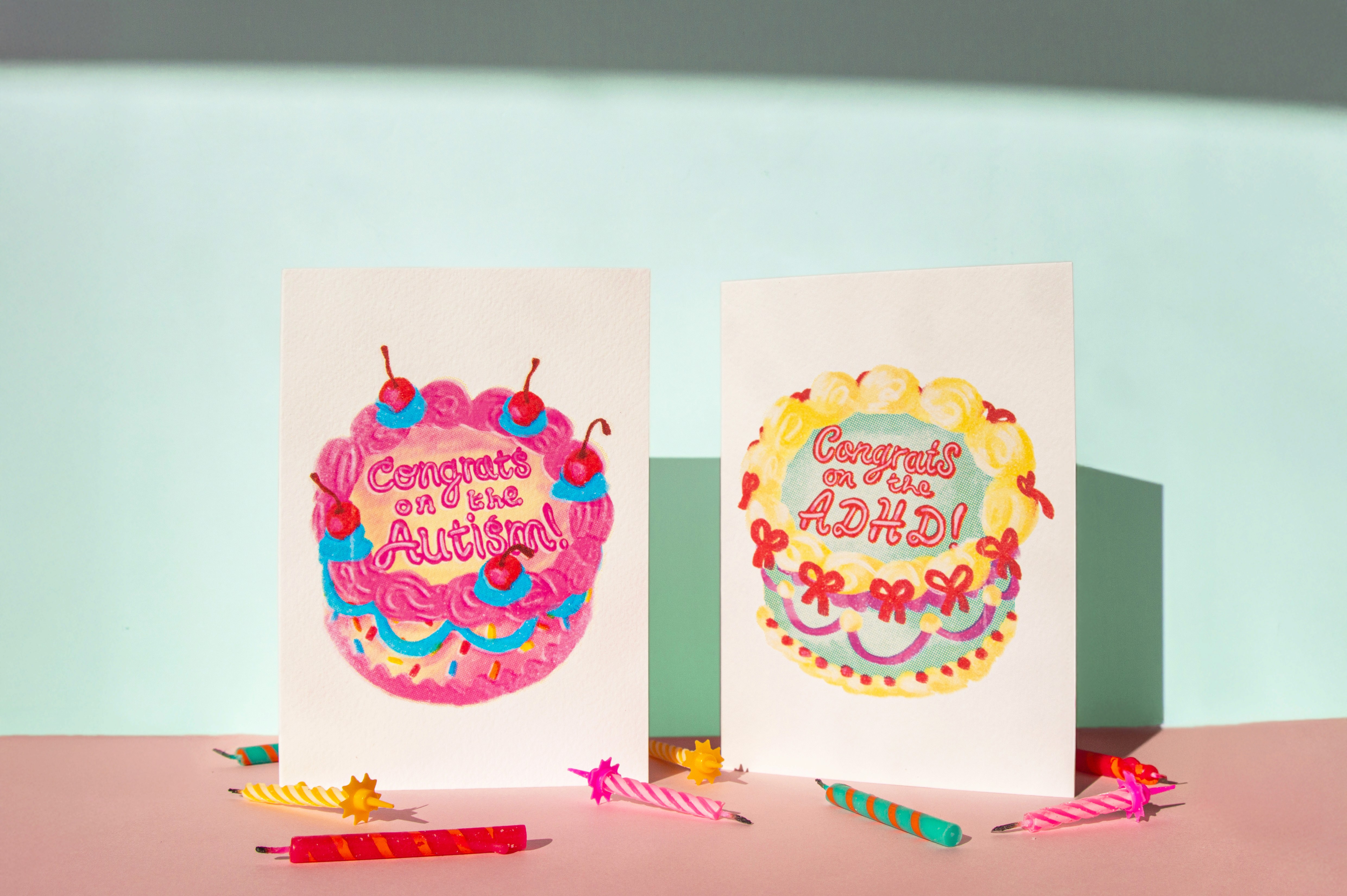 Two greetings cards stand above birthday candles. The first is a yellow and pink cake that reads "Congrats on the autism!". The second is an aqua and cream cake that reads "Congrats on the ADHD!"