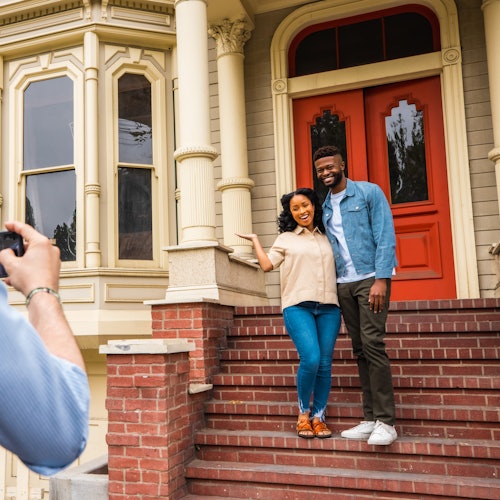 A couple poses for a photo on the steps of a house with red double doors and ornate columns while another person takes their picture.