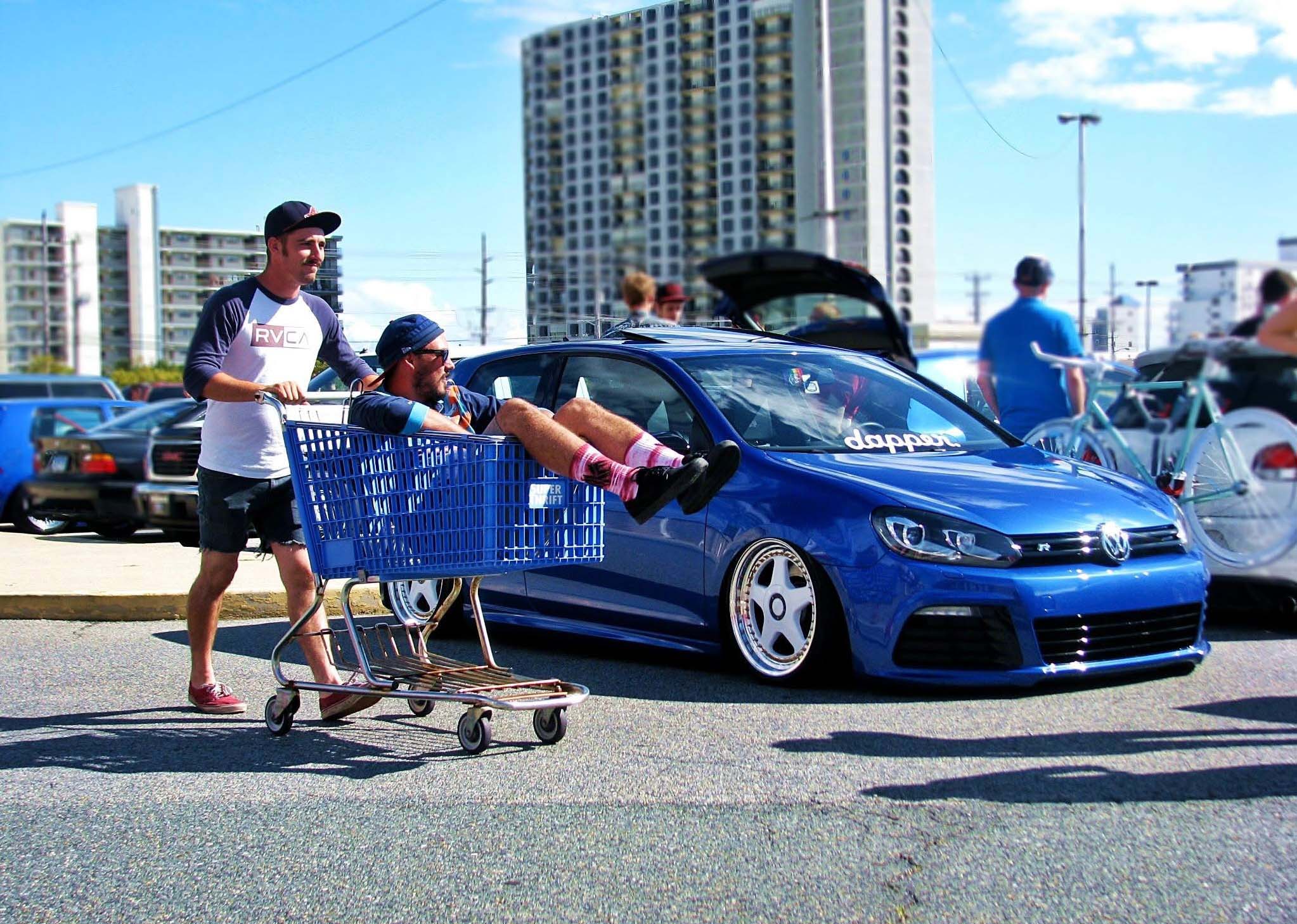 Two men with a shopping cart beside a modified blue Volkswagen Golf R at an outdoor car meet.