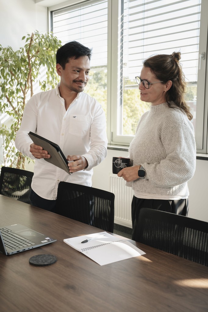 Two people discuss something over a desk, engaged and smiling, in a bright office with plants in the background.