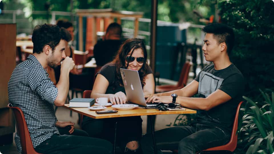 Young adults at a coffee shop, focused on their laptops and engaged in discussion, in a casual and sunny outdoor setting.