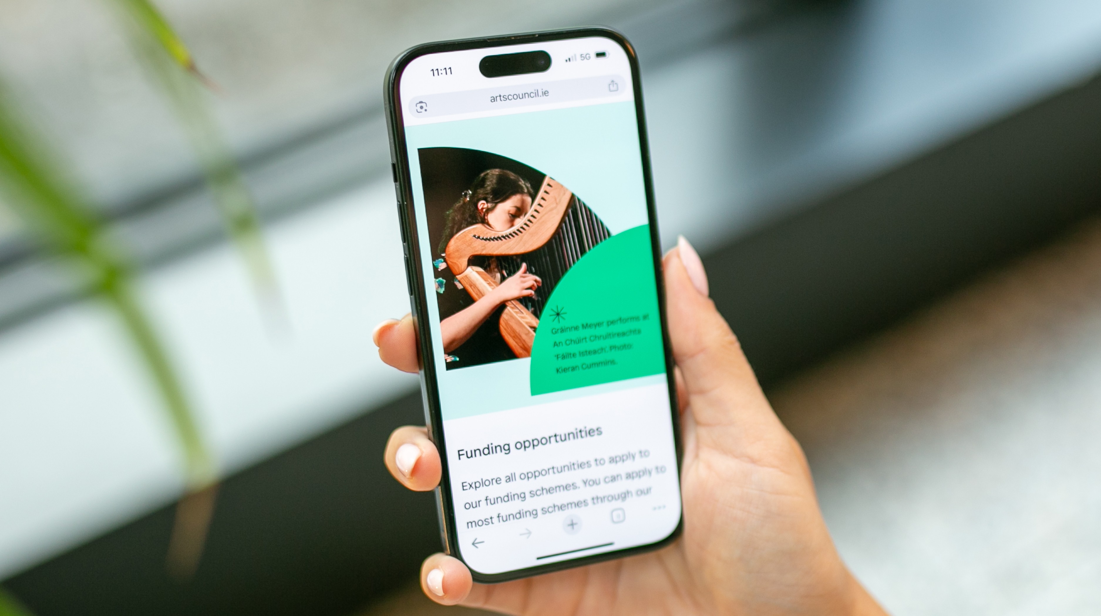 Person holding a smartphone displaying the Arts Council of Ireland website, showing a page about funding opportunities with an image of a harpist performing.
