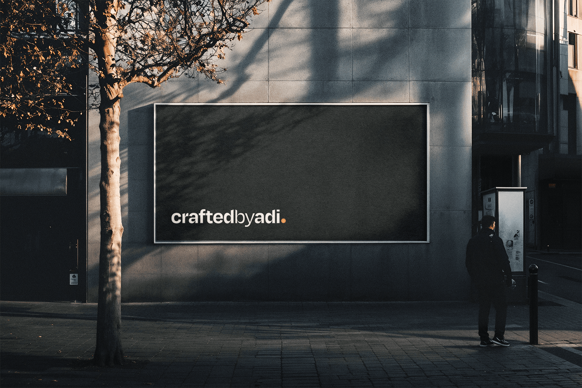 A horizontal dark billboard featuring the white 'craftedbyadi' wordmark mounted on a grey stone building facade, captured in natural late-afternoon sunlight with long shadows.