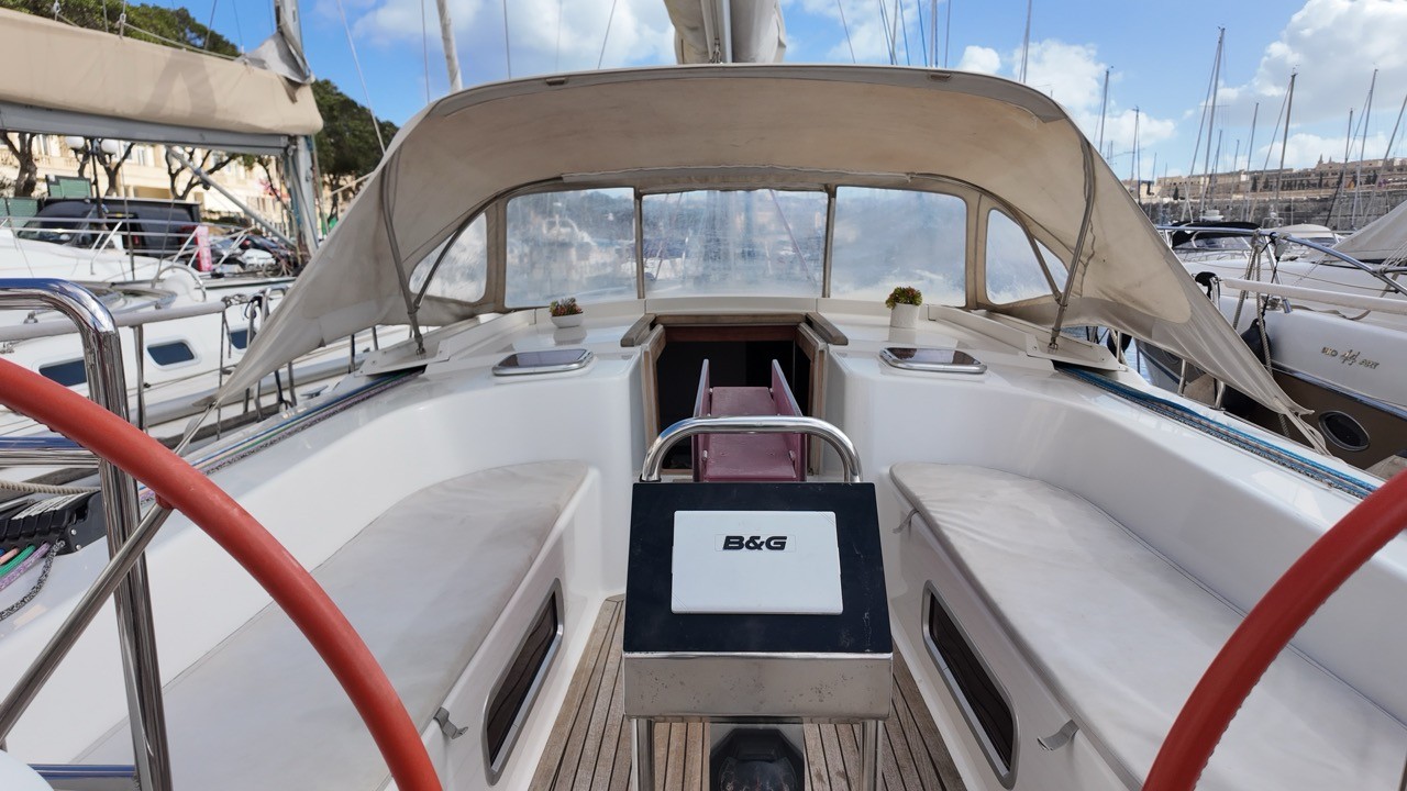Hanse 470e cockpit view looking forward under a canvas bimini with clear side screens, showing EcoDeck composite cockpit floor, B&G chartplotter on the binnacle, twin helm seats, open companionway, and marina background
