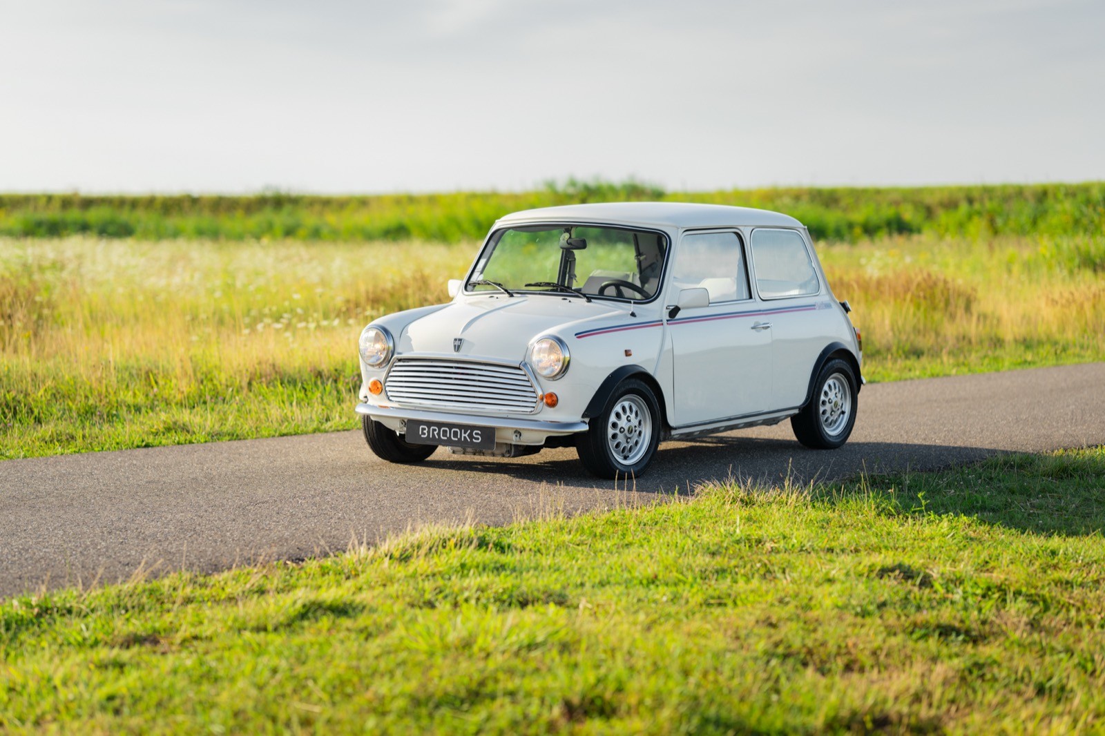 A vintage white car parked on a scenic road surrounded by green fields under a clear blue sky.