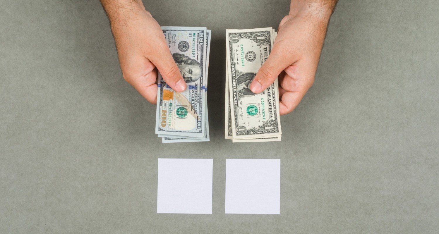 Two hands holding and comparing stacks of U.S. dollar bills, hundreds in the left hand and one-dollar notes in the right, above two blank white cards on a gray background.