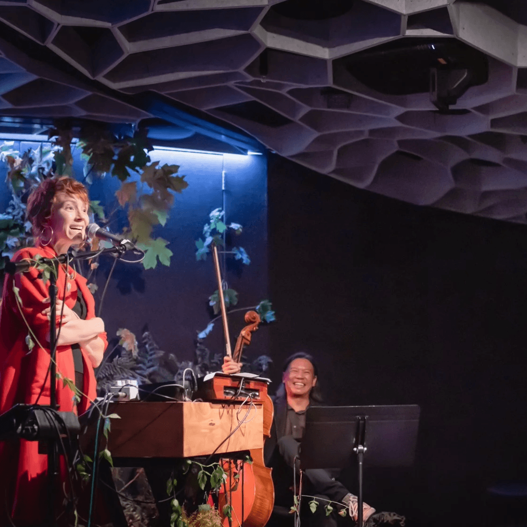 A woman stands on stage surrounded by plants an instruments, a cellist laughing in the background