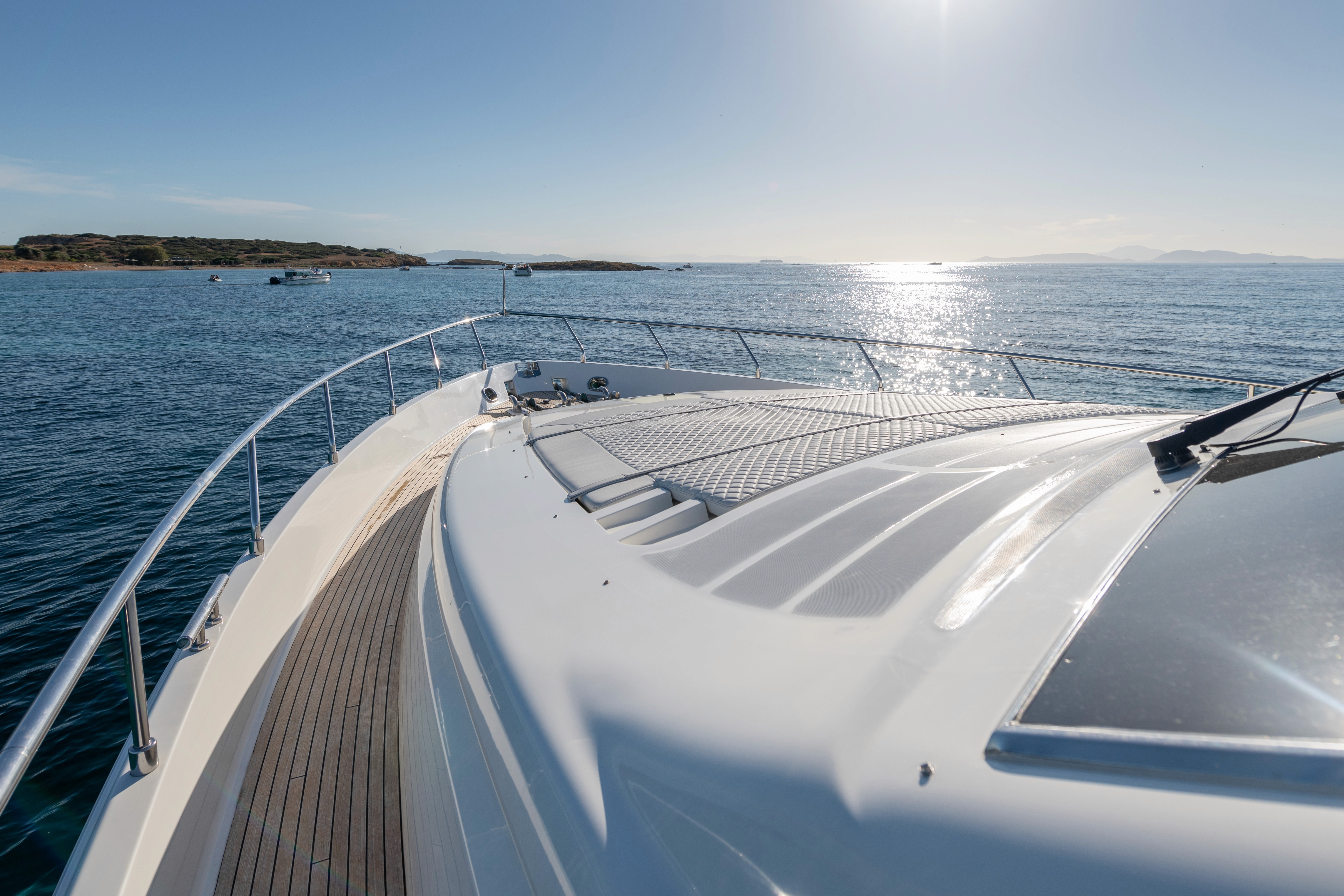 Luxury yacht deck with quilted sunpad, black towels, champagne service, and Greek island coastline in background.