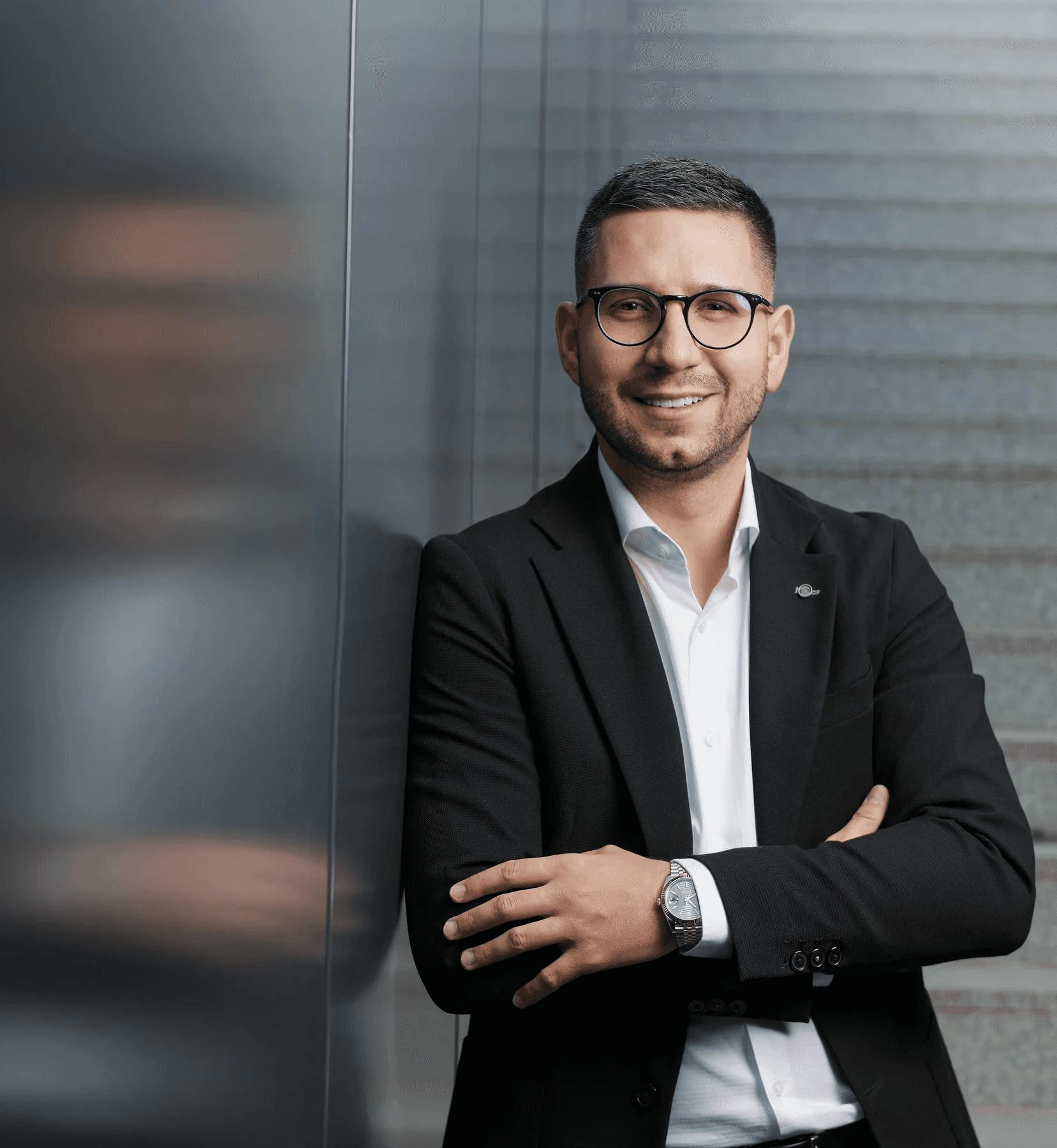 Muž s brýlemi a krýtkými vousy v bílé košili a černém saku opírající se o stěnu z ocelových panelů