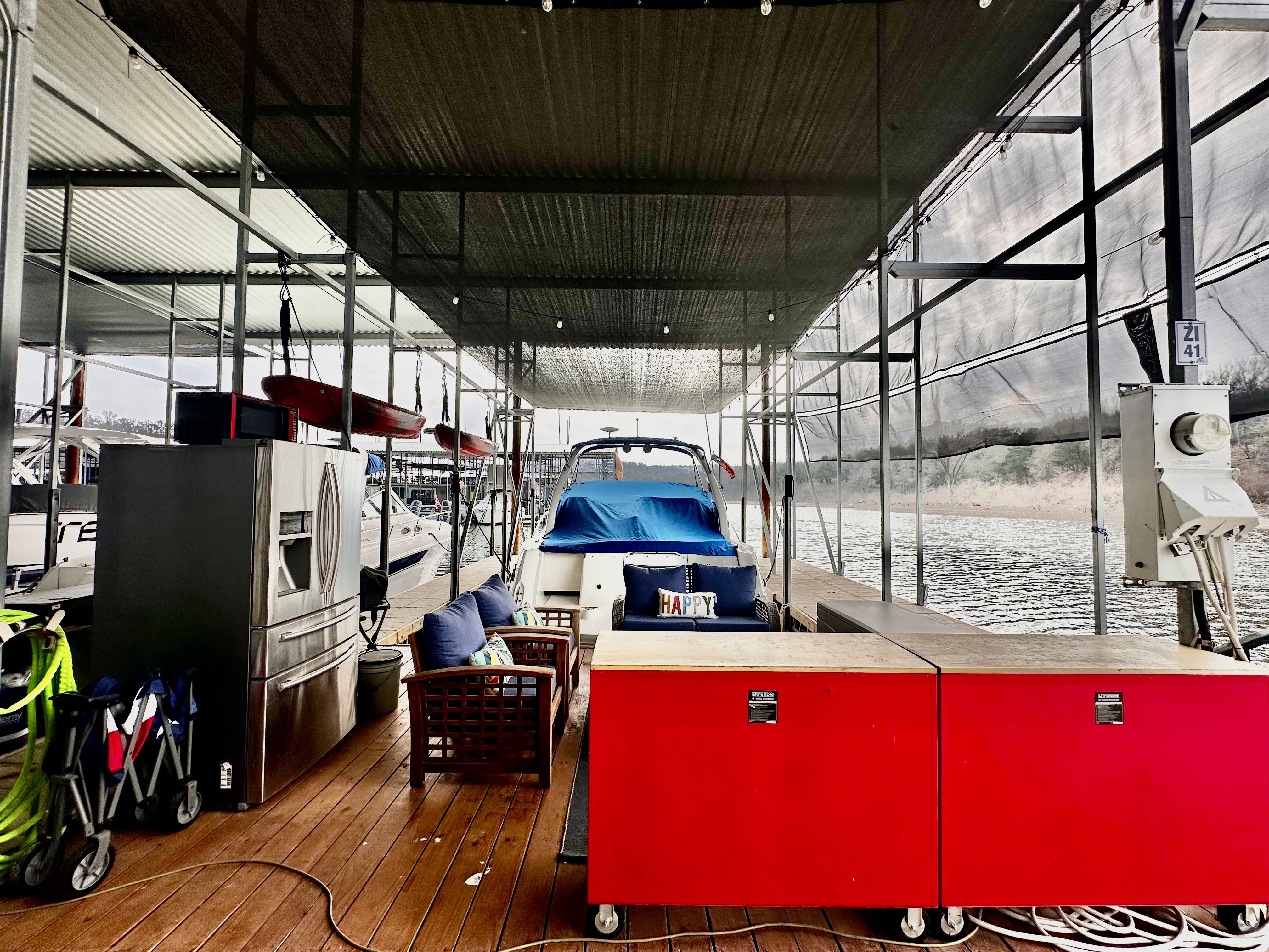 A docked boat covered with a blue tarp sits in a sheltered marina slip, surrounded by red storage units, chairs, and hanging kayaks beside a serene waterfront.