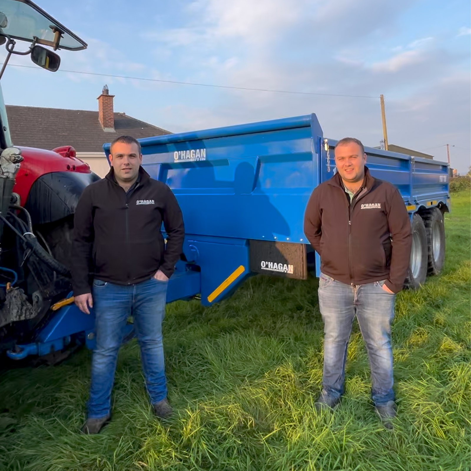 The O'Hagan Brothers smiling and standing in a field in front of a Blue O'hagan trailer pulled by a Red Massey Ferguson