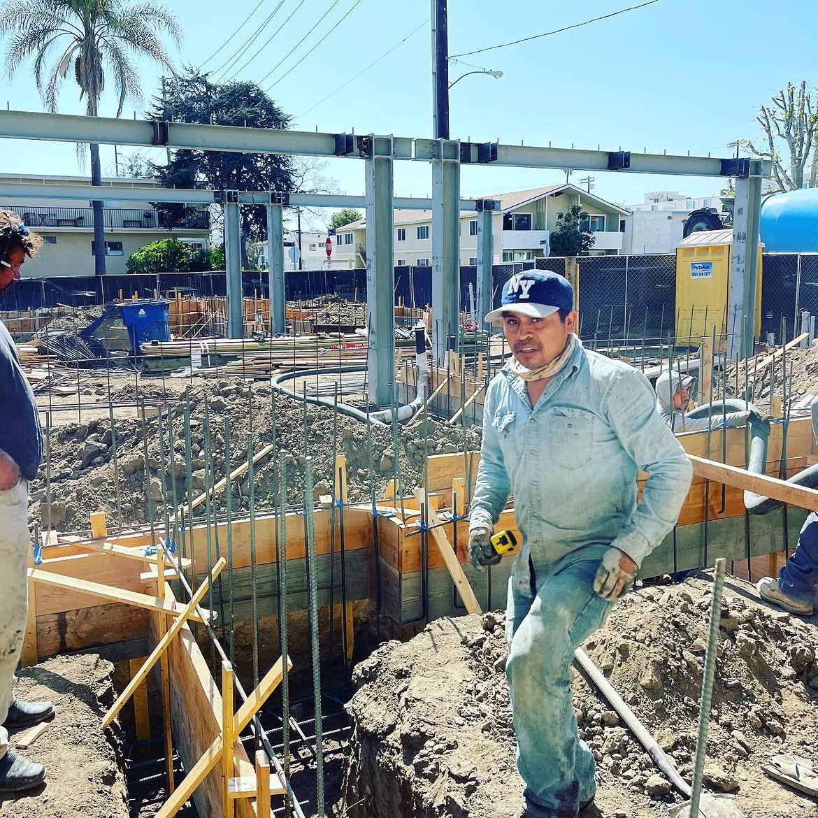 A construction worker holding a tape measure at an active building foundation site with steel beams and rebar.