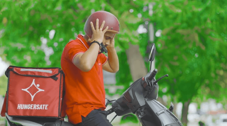 Delivery rider wearing a helmet beside a Hungersate delivery bag in an ad film.