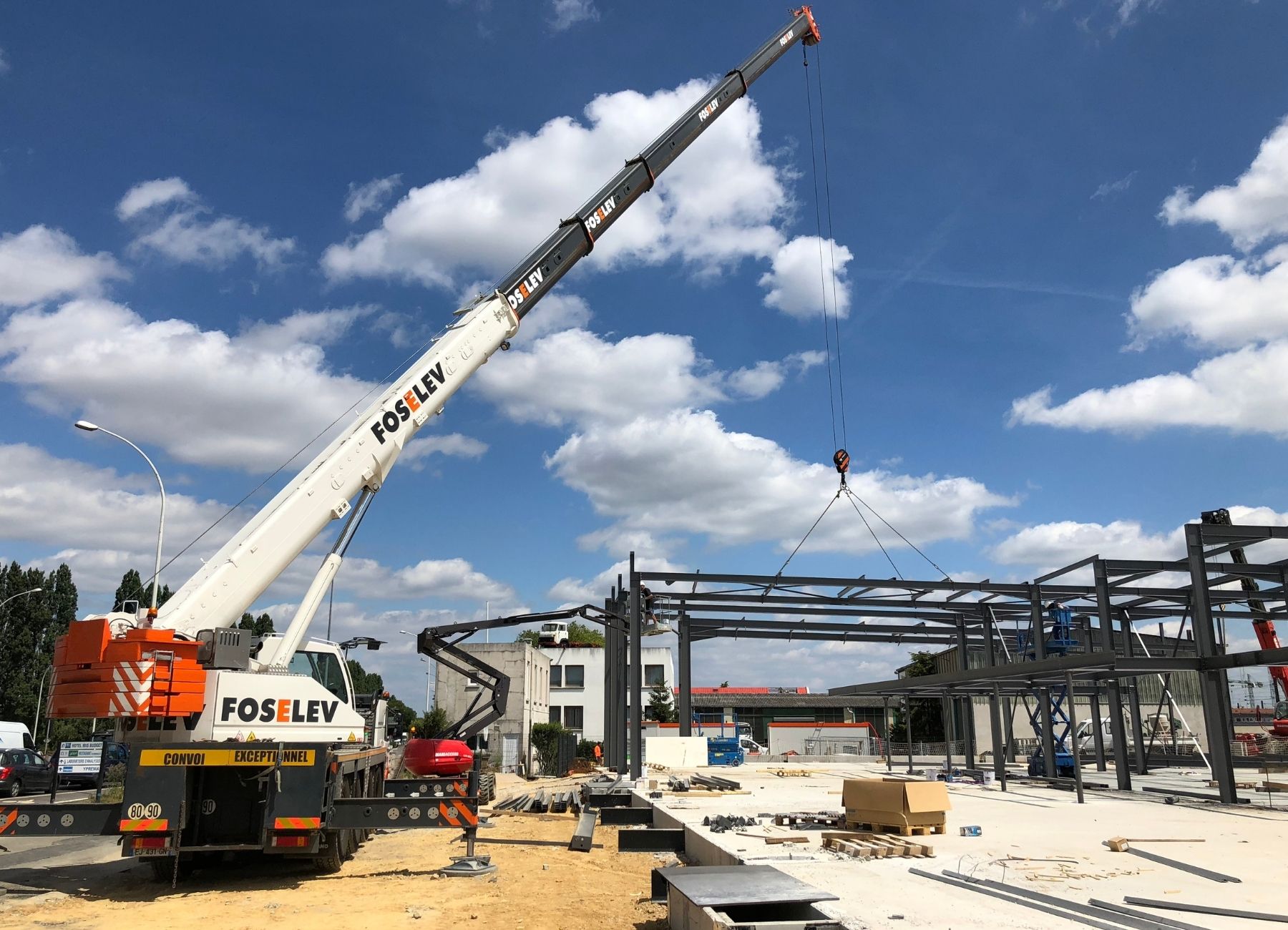 Grue mobile soulevant des éléments de charpente métallique sur chantier industriel.