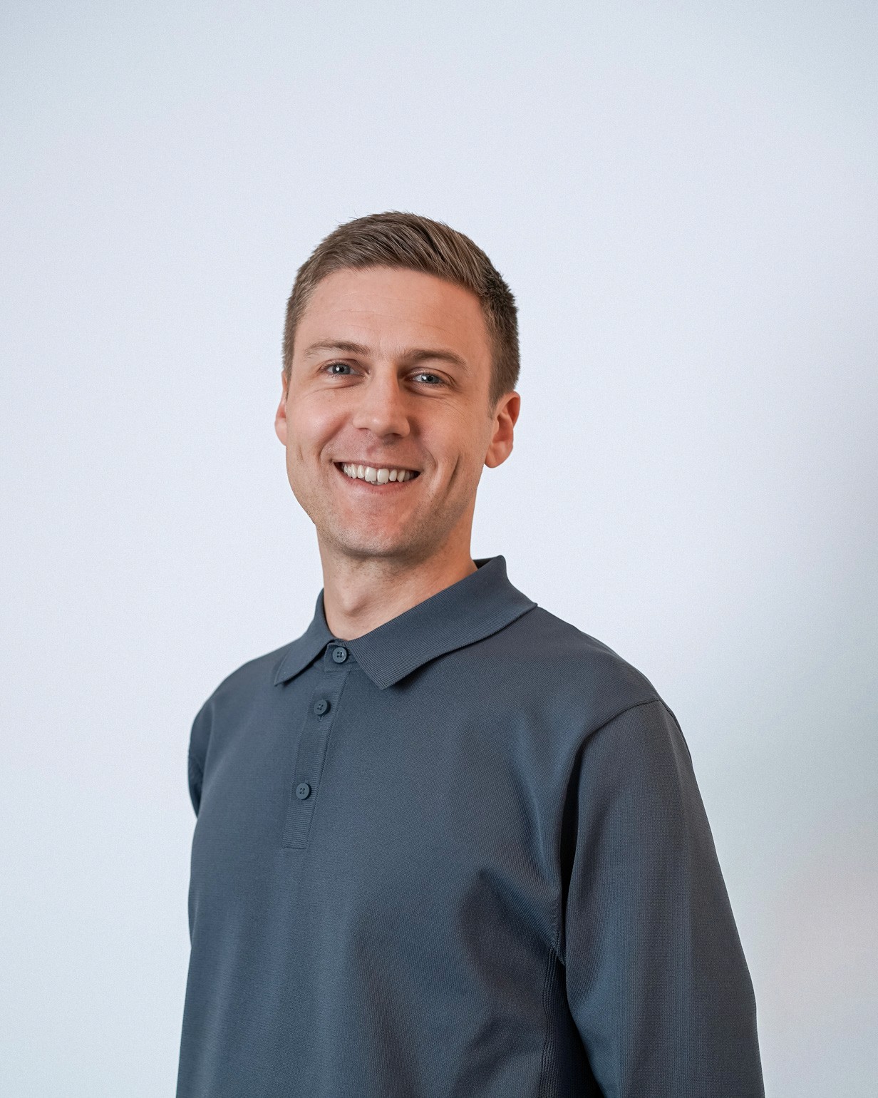 Man wearing long sleeved grey collared shirt and smiling in front of purple background.