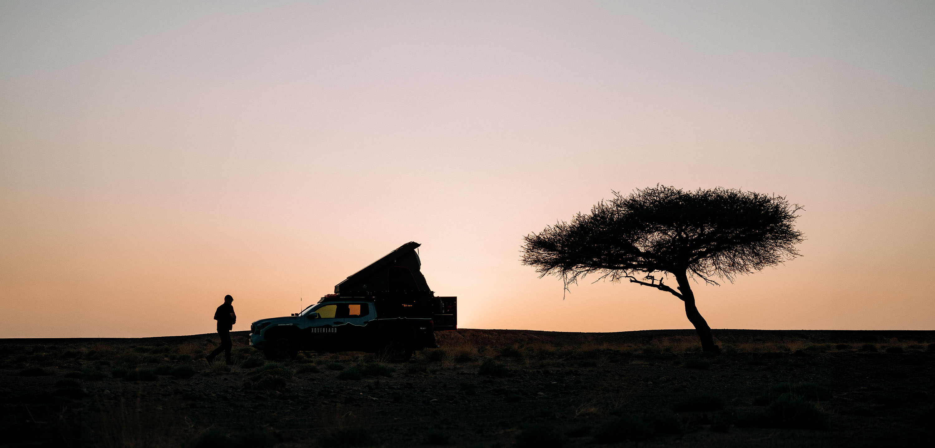 Silhouette of an overland vehicle with a rooftop tent and a person standing beside it on a ridge at sunset, with a lone tree to the right against a clear sky.