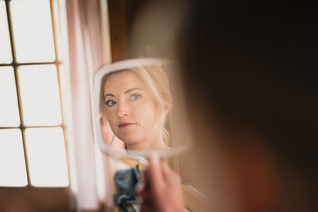 A close-up reflection in a small hand mirror showing a bride with blonde hair in a soft updo, looking at her finished makeup with a neutral, thoughtful expression.