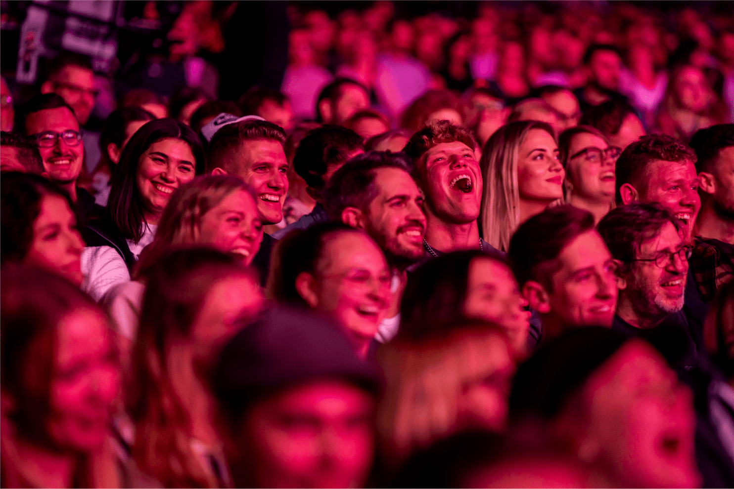 A crowd of people enjoying an event, illuminated by vibrant pink lights, with many smiling and engaged.