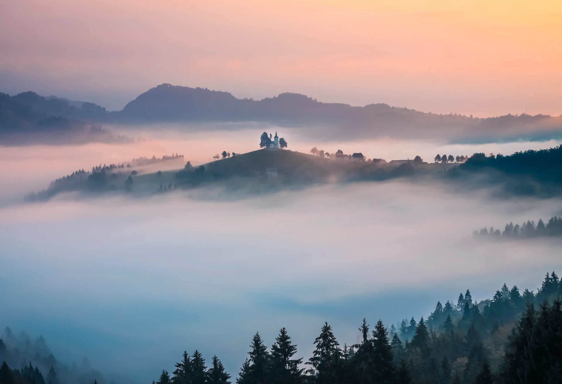 A panoramic view of the Church of St. Thomas (Sveti Tomaž) perched on a small hill in Slovenia, surrounded by a thick sea of blue-tinted morning mist.