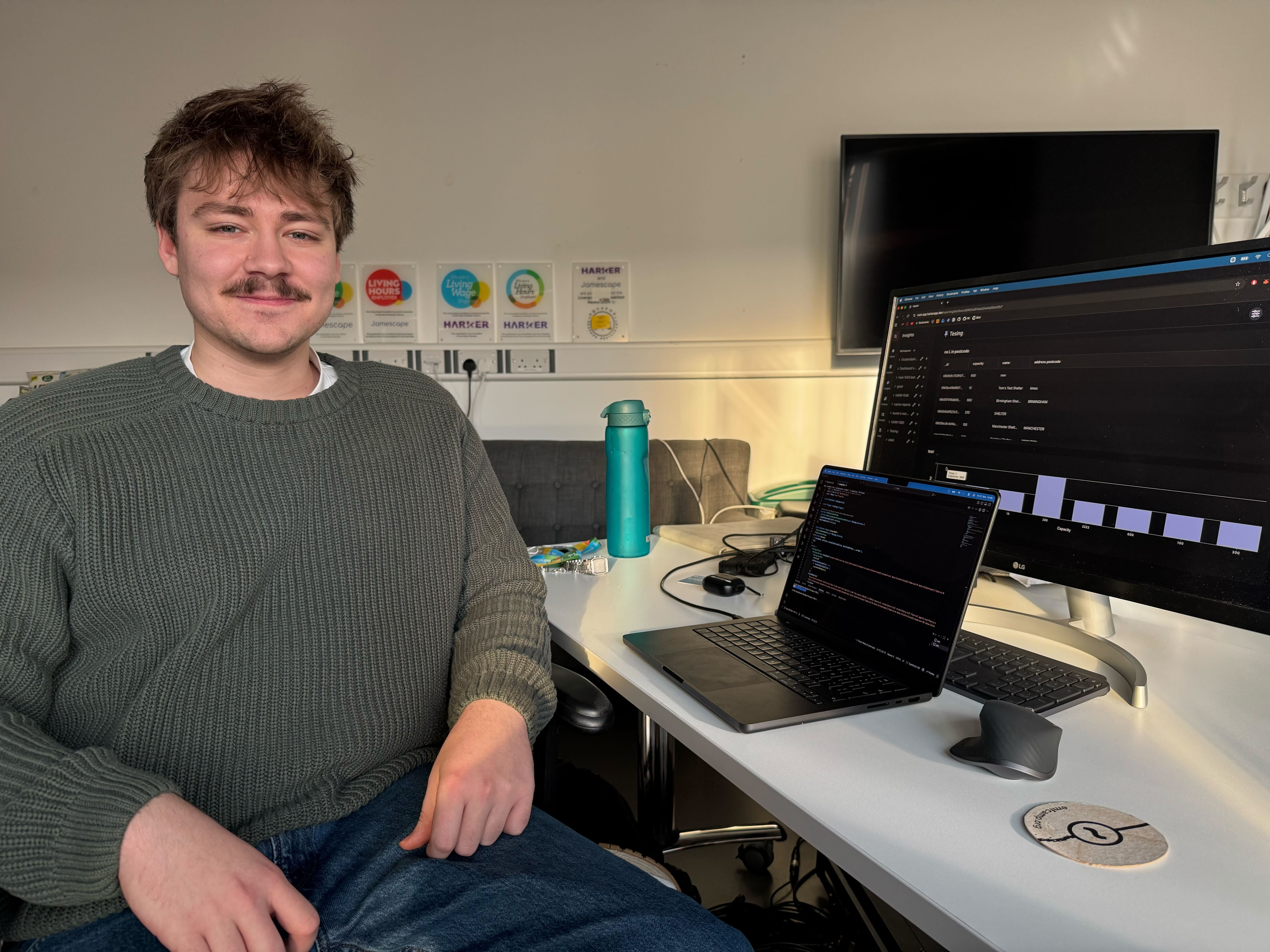 Josh sat at his desk in the office, smiling at the camera. His monitor shows a test screenshot of Harker.