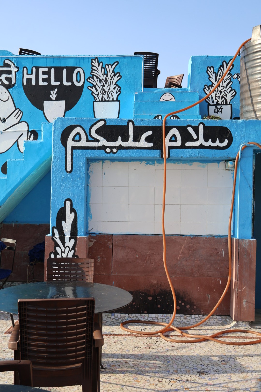Mural painting showing potted plants, joyful cartoon characters and the word ‘Hello, नमस्ते, سلام عليكم’ painted on a blue building terrace with restaurant tables visible.