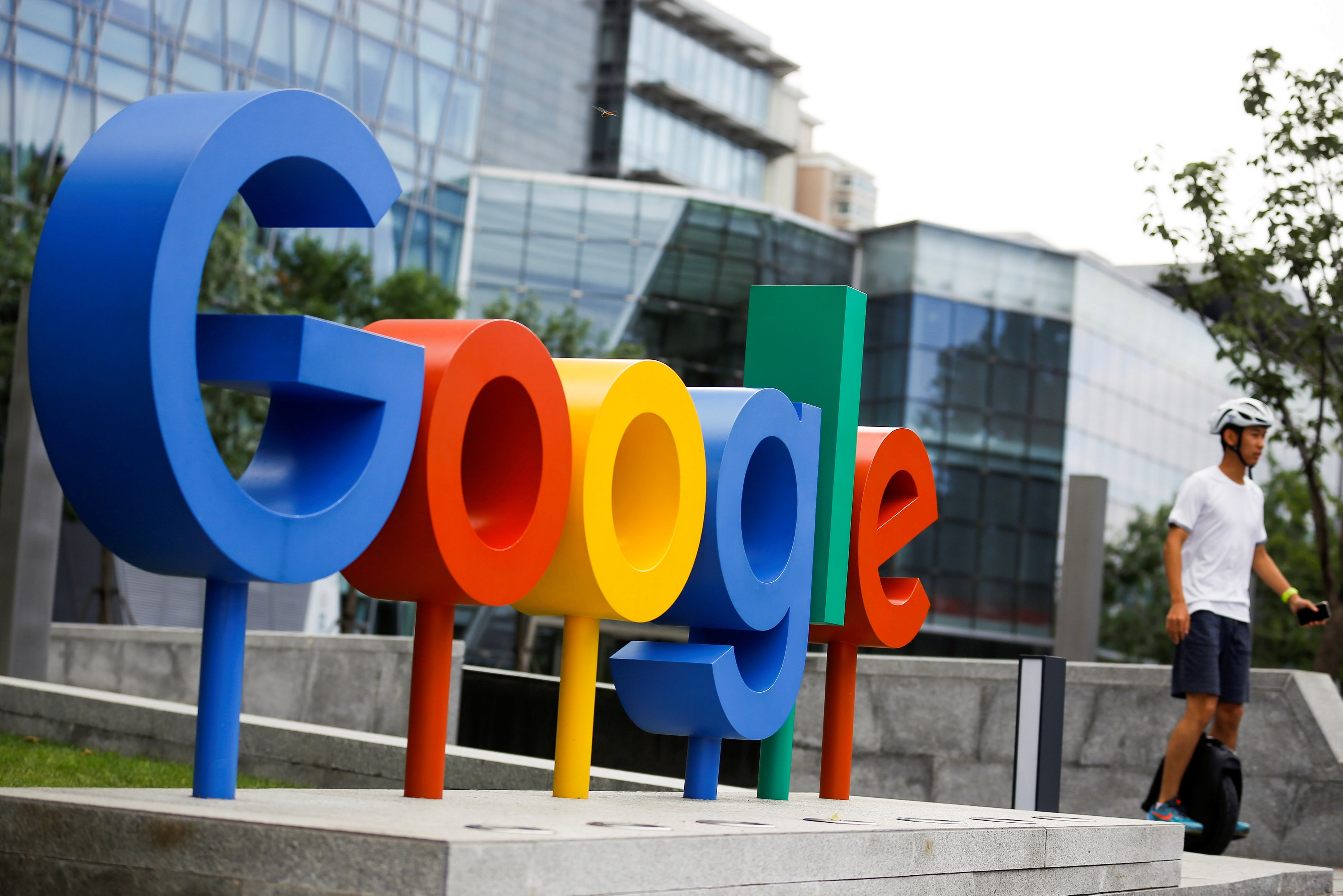 Colorful Google logo displayed outdoors in front of a modern glass building, with a person walking by, emphasizing Google's brand presence in an urban setting.