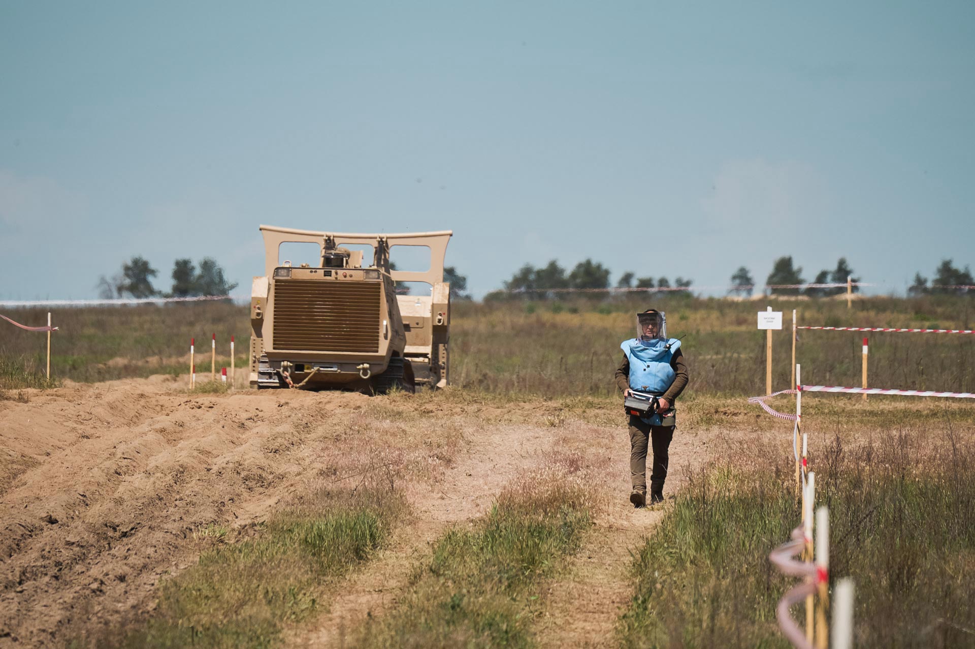 GCS demining operator in blue protective vest and visor helmet operating alongside a GCS-200 demining machine on a mine-contaminated field during humanitarian demining operations.