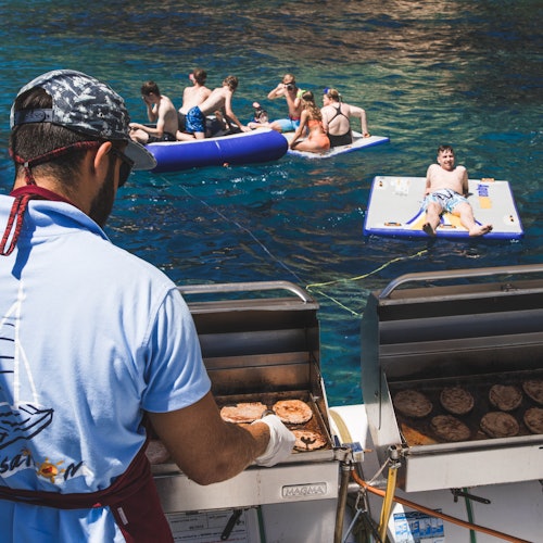 Un homme grille des hamburgers sur un bateau pendant que des personnes en maillots de bain se détendent sur des plateformes flottantes dans une eau bleue claire à proximité.