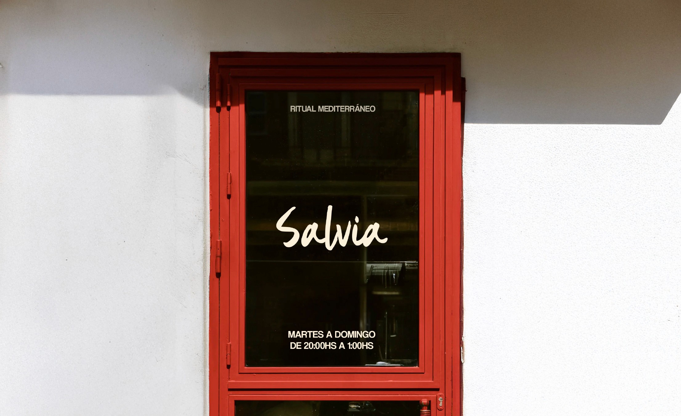A window with a red frame displays the words "Salvia" in white script, along with Spanish text detailing business hours, set against a minimalist white wall, embodying a modern Mediterranean theme.