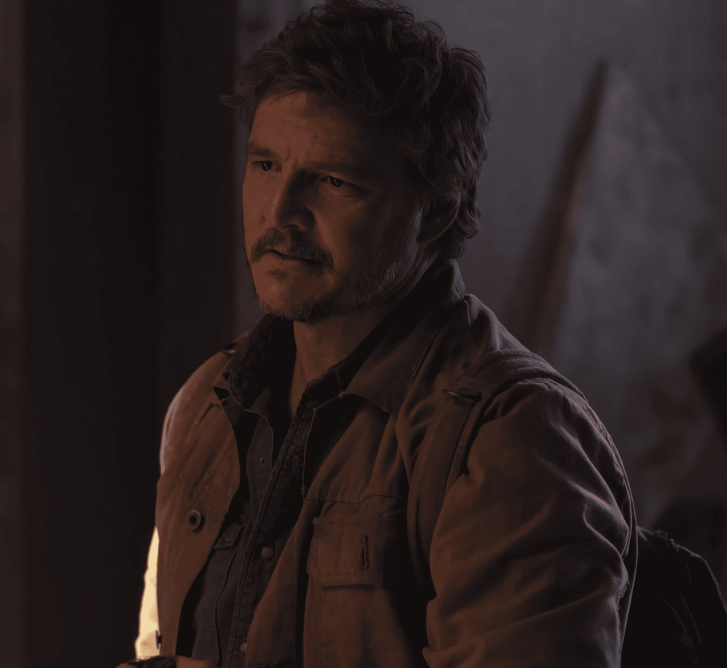 Pedro Pascal as Joel in 'The Last of Us', wearing a jacket and looking serious in dim light.