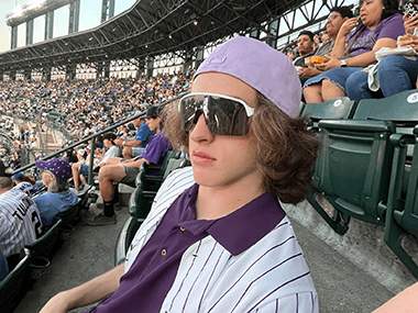 Cooper side-eyes the camera while sitting in the stands at a Rockies game. He is dressed up in purple rockies gear.