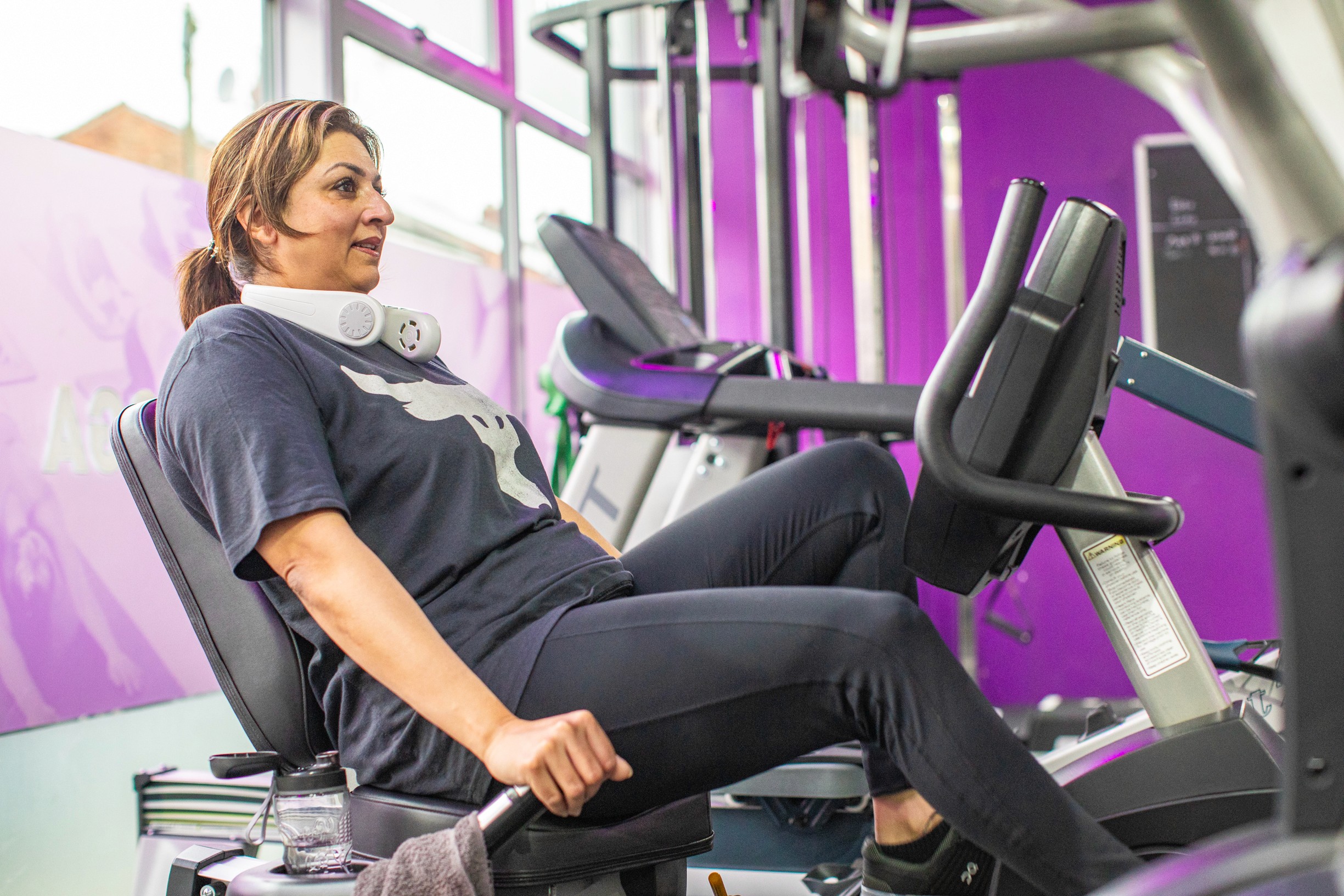 A woman works out on a recumbent exercise bike in a gym. She wears headphones around her neck, focusing intently. The room is brightly lit and purple-themed.