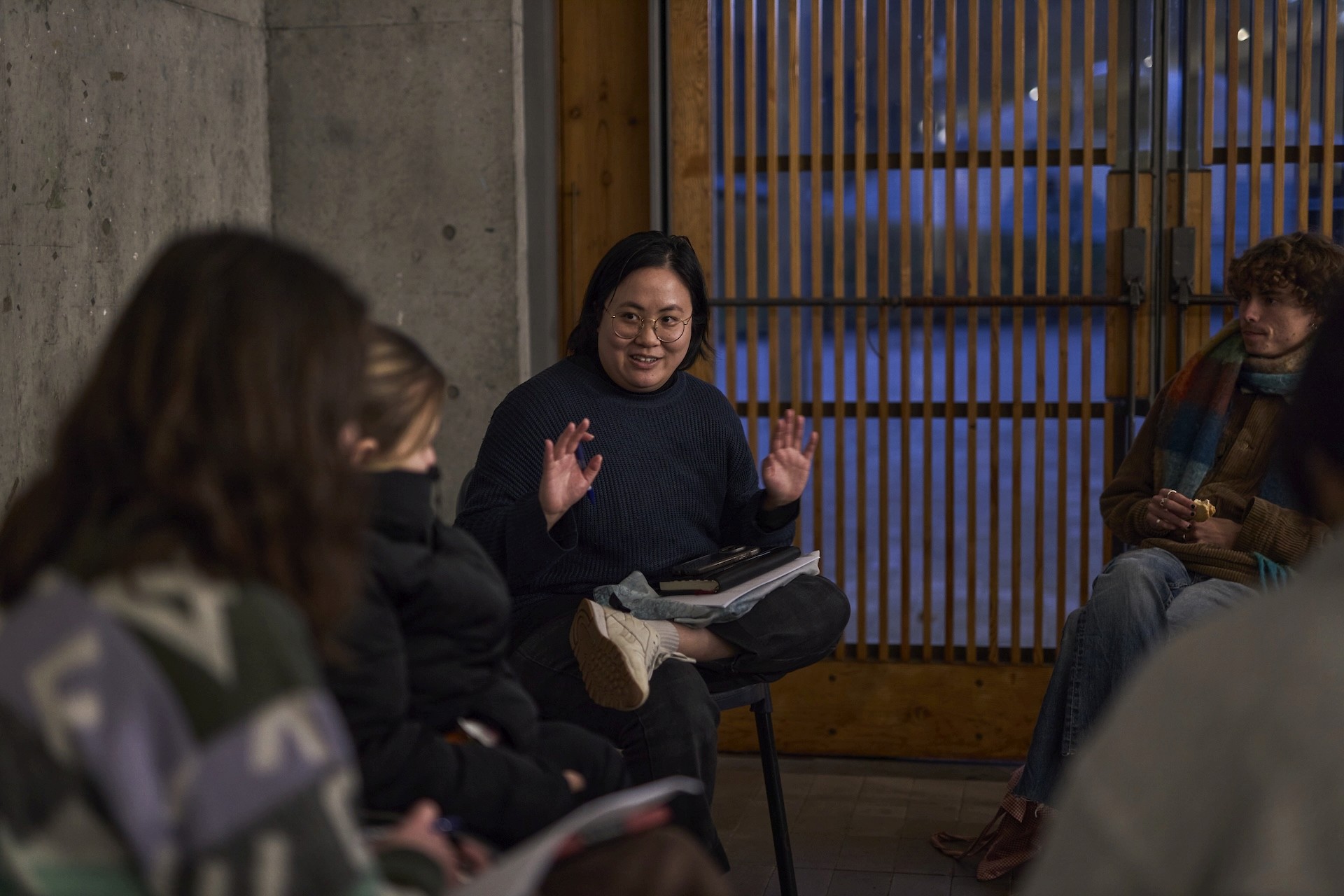 A person sits cross-legged on a chair in a small group discussion, speaking with their hands raised mid-gesture. They wear glasses and a dark jumper, with notebooks on their lap. Several people sit around them listening. The setting appears informal, with concrete walls and wooden slats behind them, suggesting a workshop or creative discussion space in the evening.