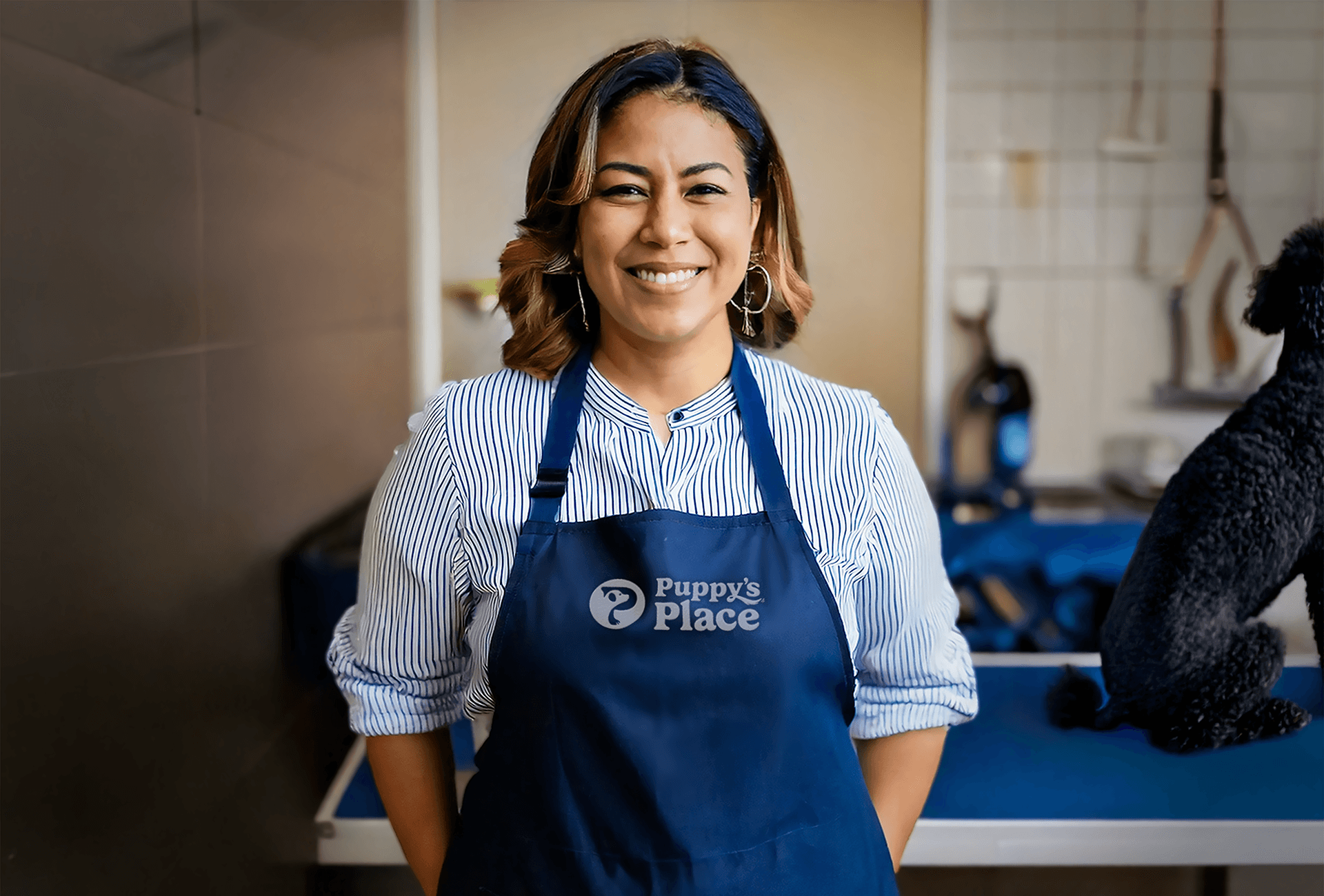 A smiling groomer wearing an apron with the Puppy's Place logo embroidered on the front