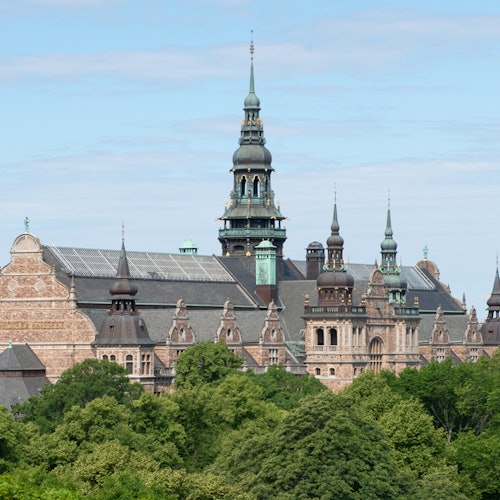Történelmi épület több toronnyal és díszes részletekkel, buja zöld fák övezik, részben felhős ég alatt.