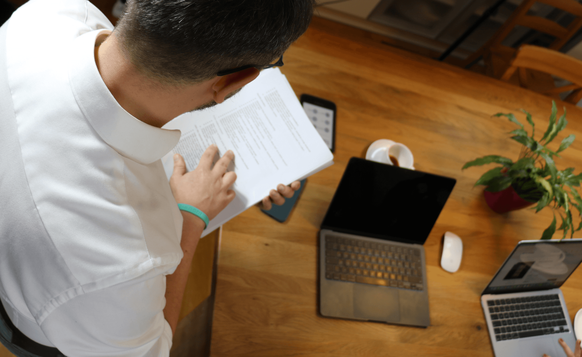 Osoba čtoucí notebook, zatímco sedí u dřevěného stolu s notebookem a malou rostlinou v květináči.