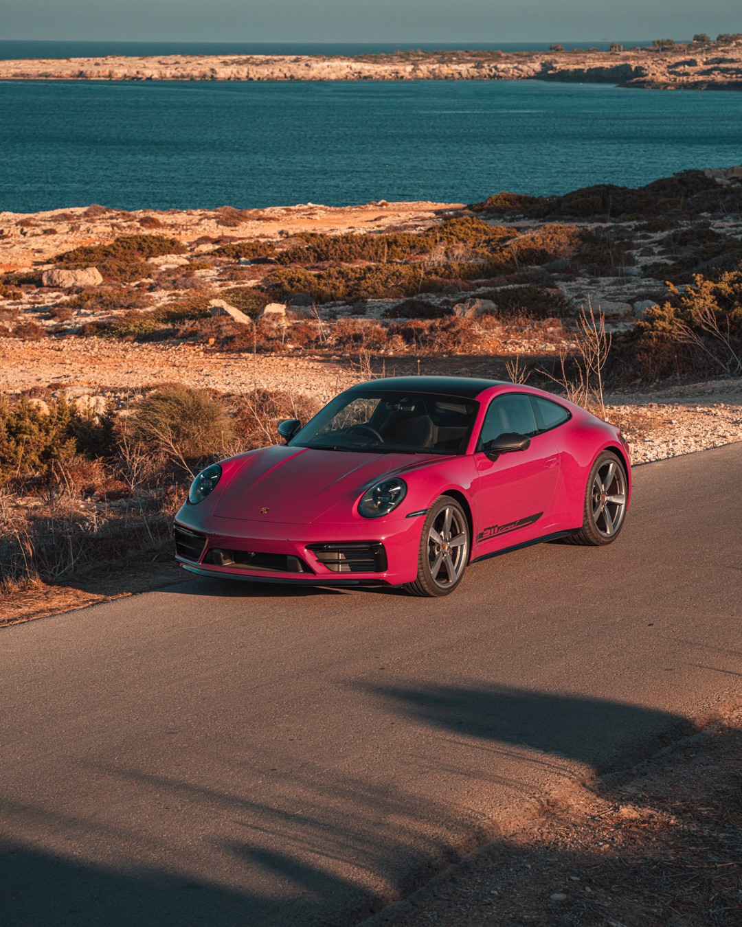 Ruby Star Porsche 911 Carrera T (992) front three-quarter on a coastal road at golden hour, gold "911 Carrera T" decal visible, with rocky scrubland and the blue sea behind, in Cyprus.