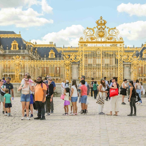Menschen versammeln sich vor einem großen, kunstvoll gestalteten goldenen Tor mit einem komplexen Design, das vor einem historischen Palast unter einem teilweise bewölkten Himmel steht.