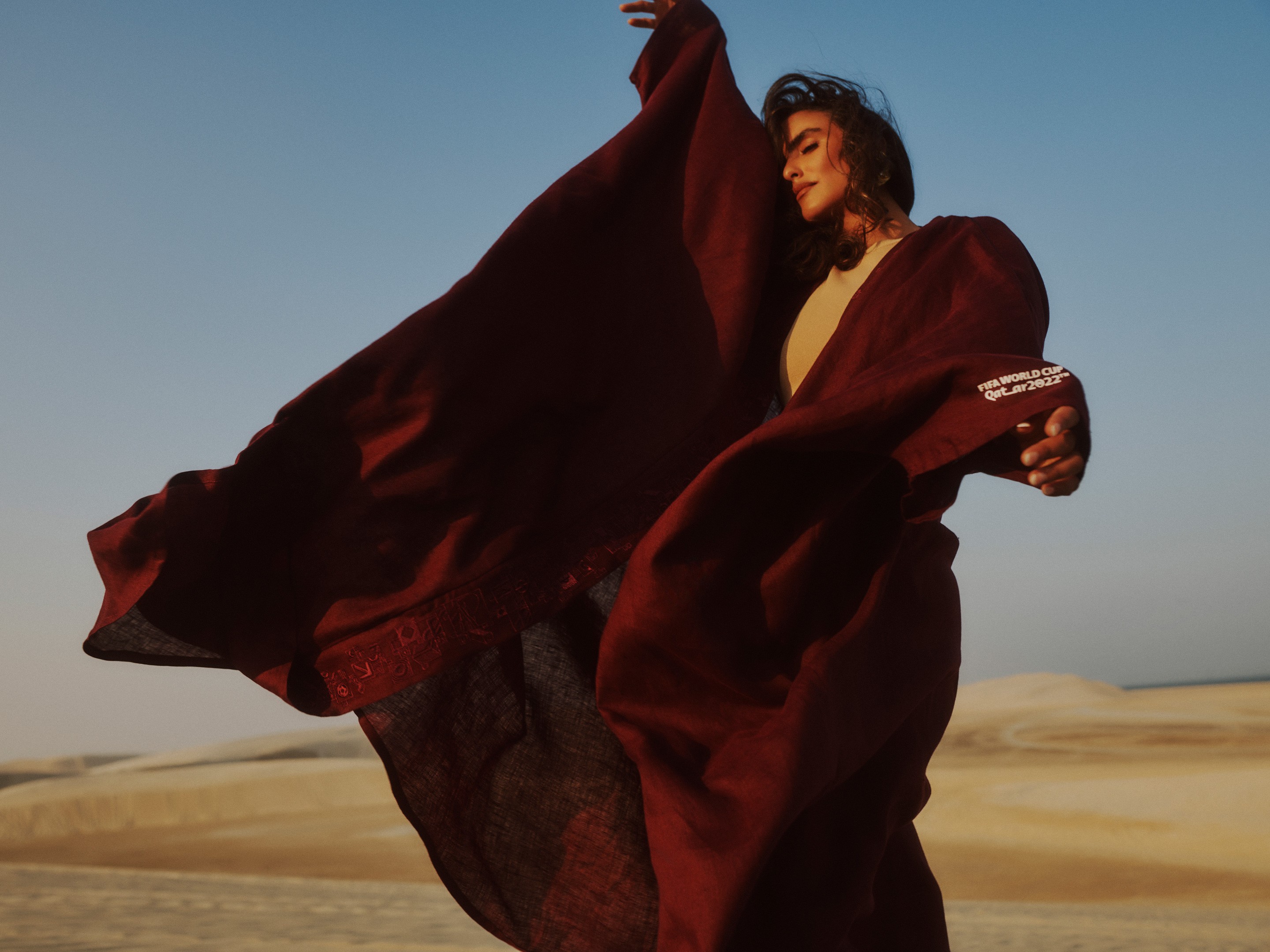 A model in a flowing maroon abaya, the fabric caught mid-billow across sunlit desert dunes.