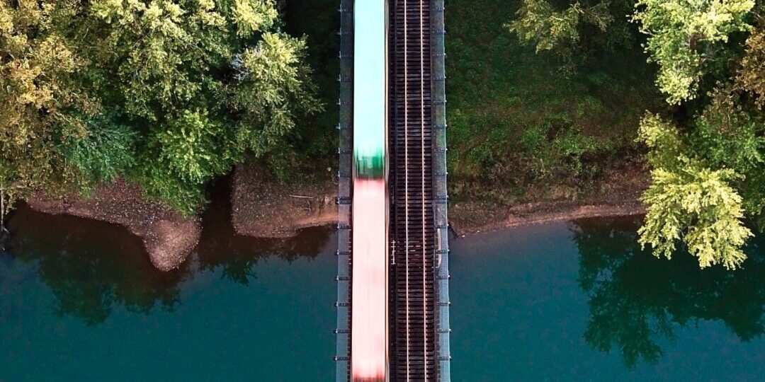 Aerial view of a colorful train crossing a narrow railroad bridge over a tranquil river, surrounded by lush green trees on both sides.