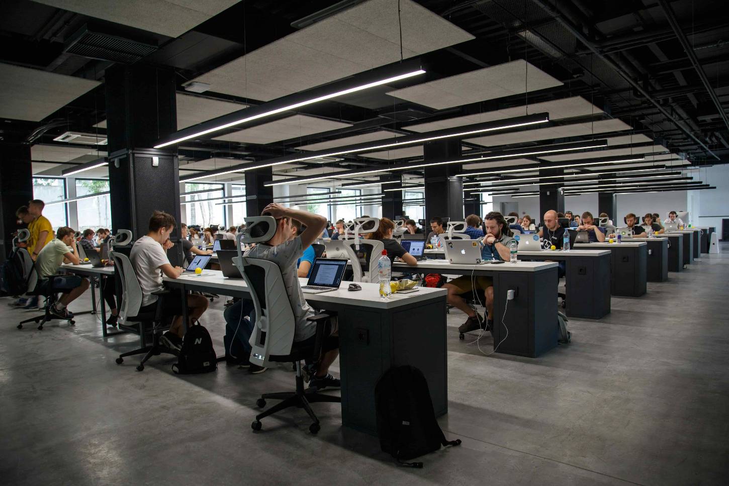 Ein weitläufiges, modernes Großraumbüro mit industriellem Design. Zahlreiche junge Menschen sitzen konzentriert an langen, weißen Schreibtischreihen und arbeiten an ihren Laptops. Die Einrichtung ist in schlichtem Grau und Weiß gehalten, ergänzt durch markante schwarze Säulen und hängende LED-Lichtleisten an der dunklen Decke.