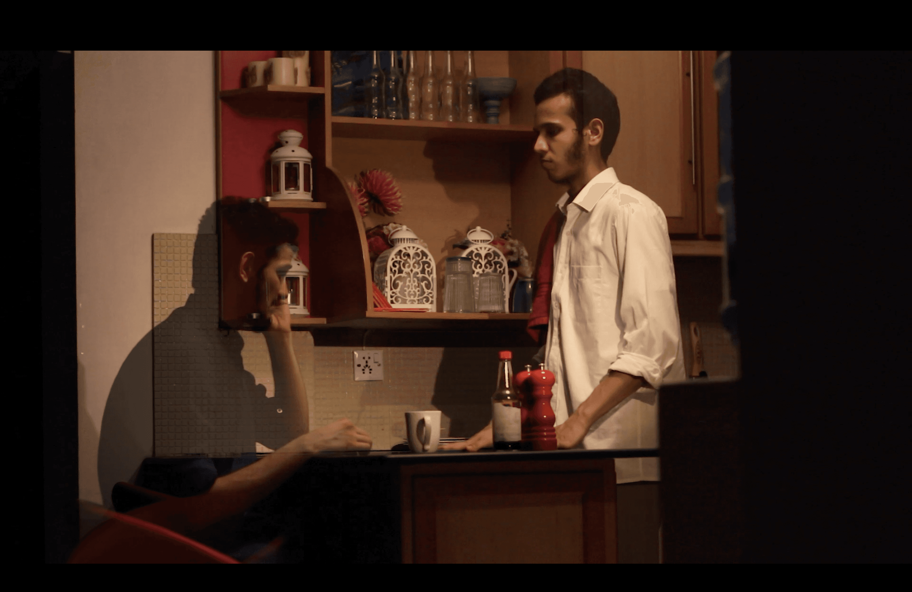 a man sitting while another man stands in front of him they are in a kitchen and the man sitting is translucent