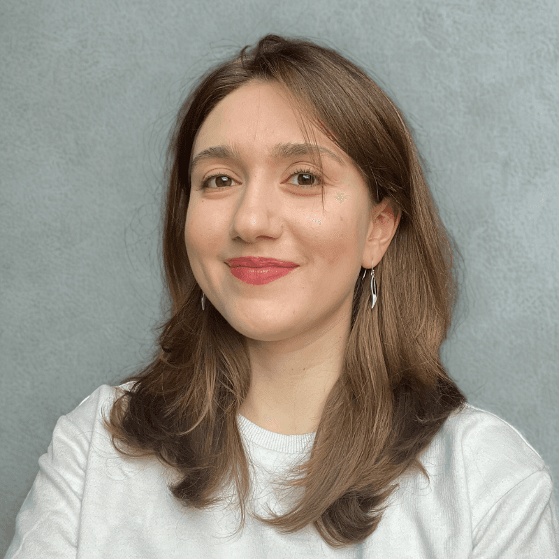 A young person with curly hair smiles at the camera while sitting in front of a laptop against a dark background.