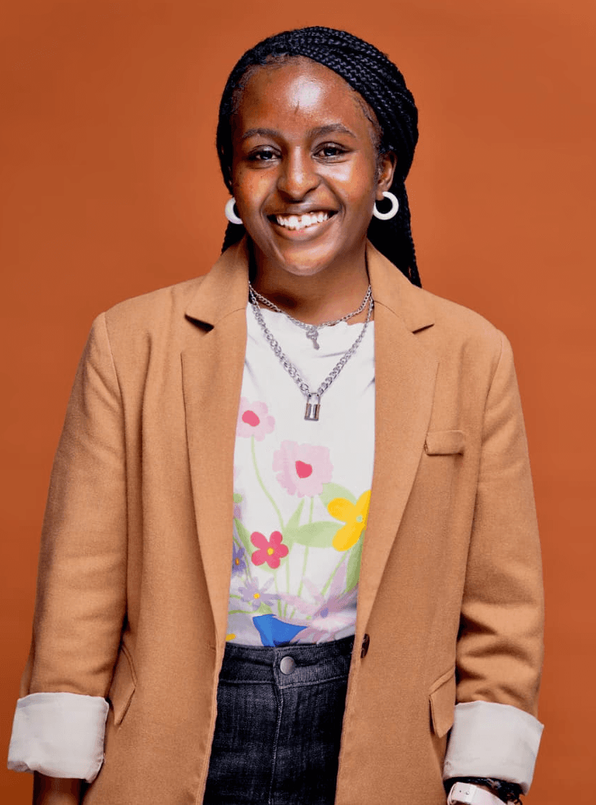 A smiling person with curly hair, wearing a leather jacket, stands in a cozy indoor setting.