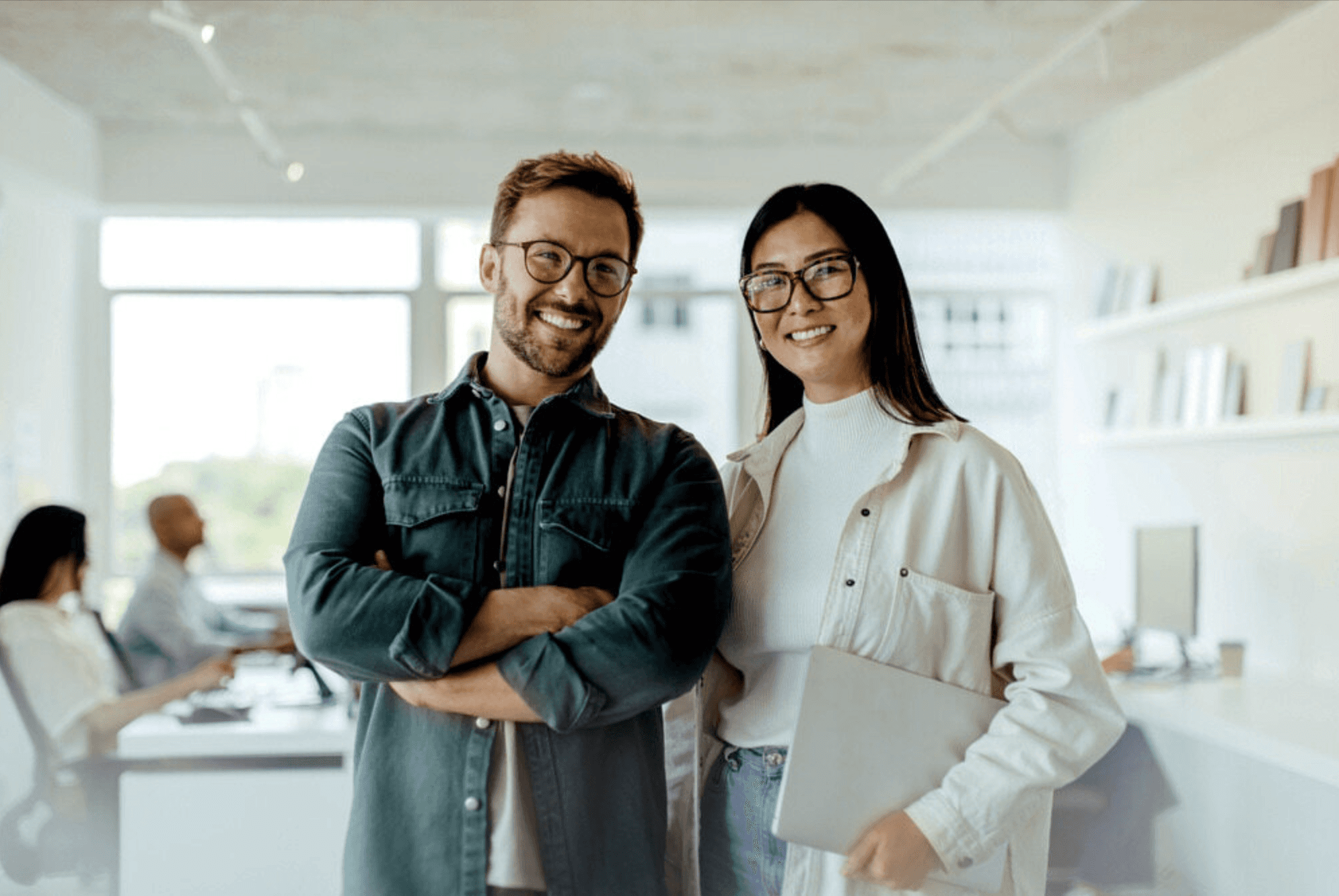 A smiling man with his arms crossed and a woman holding a laptop stand together in a bright, modern office space. Both are wearing glasses and casual work attire, with other colleagues working at desks in the blurred background.